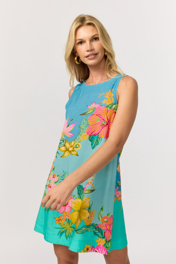 A woman wearing a sleeveless turquoise shift mini dress with a pink, yellow and green tropical floral print fading to aqua, against a light gray studio background.