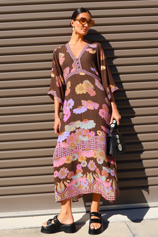 A woman stands against a brown corrugated metal wall wearing a brown kaftan dress with a colorful floral and geometric print in pink, purple, yellow, and blue. She is also wearing black slide sandals and holding a small black handbag.