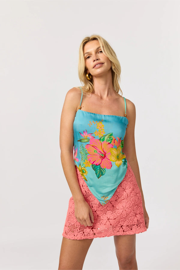 A woman modeling a two-piece tropical print set consisting of a turquoise and pink floral camisole top and a matching coral pink lace skirt, against a light gray studio background.
